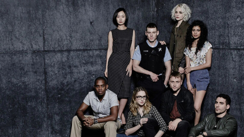 studio photo of eight people wearing dark or neutral clothing against a dark grey backdrop