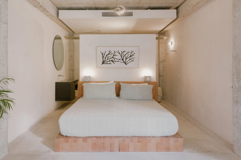 Low wooden platform kingsize bed in center of small white bedroom with circular mirror on the left wall, framed artwork over the bed covered in white bedding.