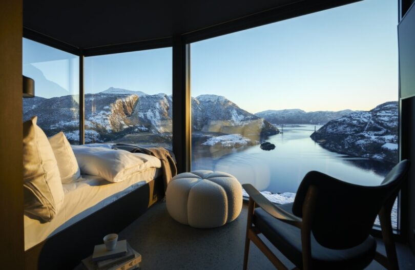 A majestic panoramic view of the nearby fjord located below the series of cabins from the glass facade covered bedroom. Interior is furnished with armchair, bed and pouf.