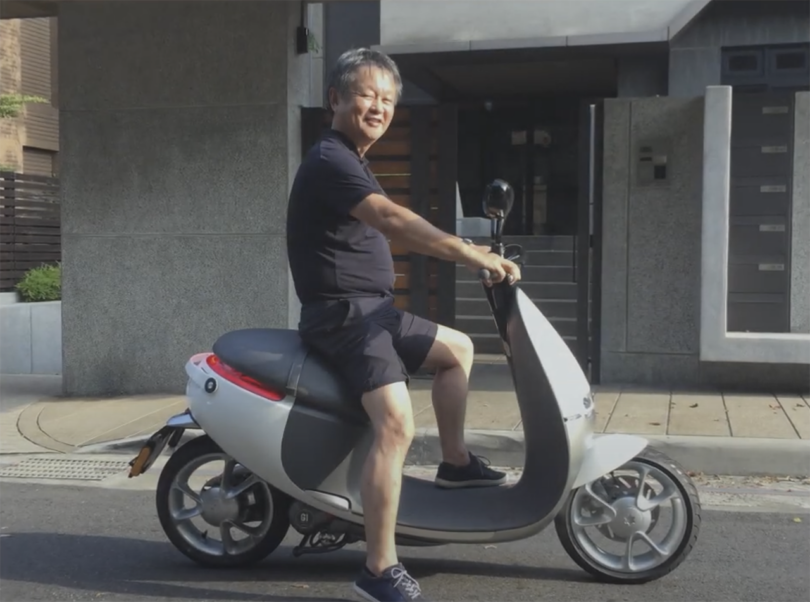 Japanese designer Naoto Fukasawa in black t-shirt and shorts seated on a Gogoro electric scooter posing on a city street smiling.