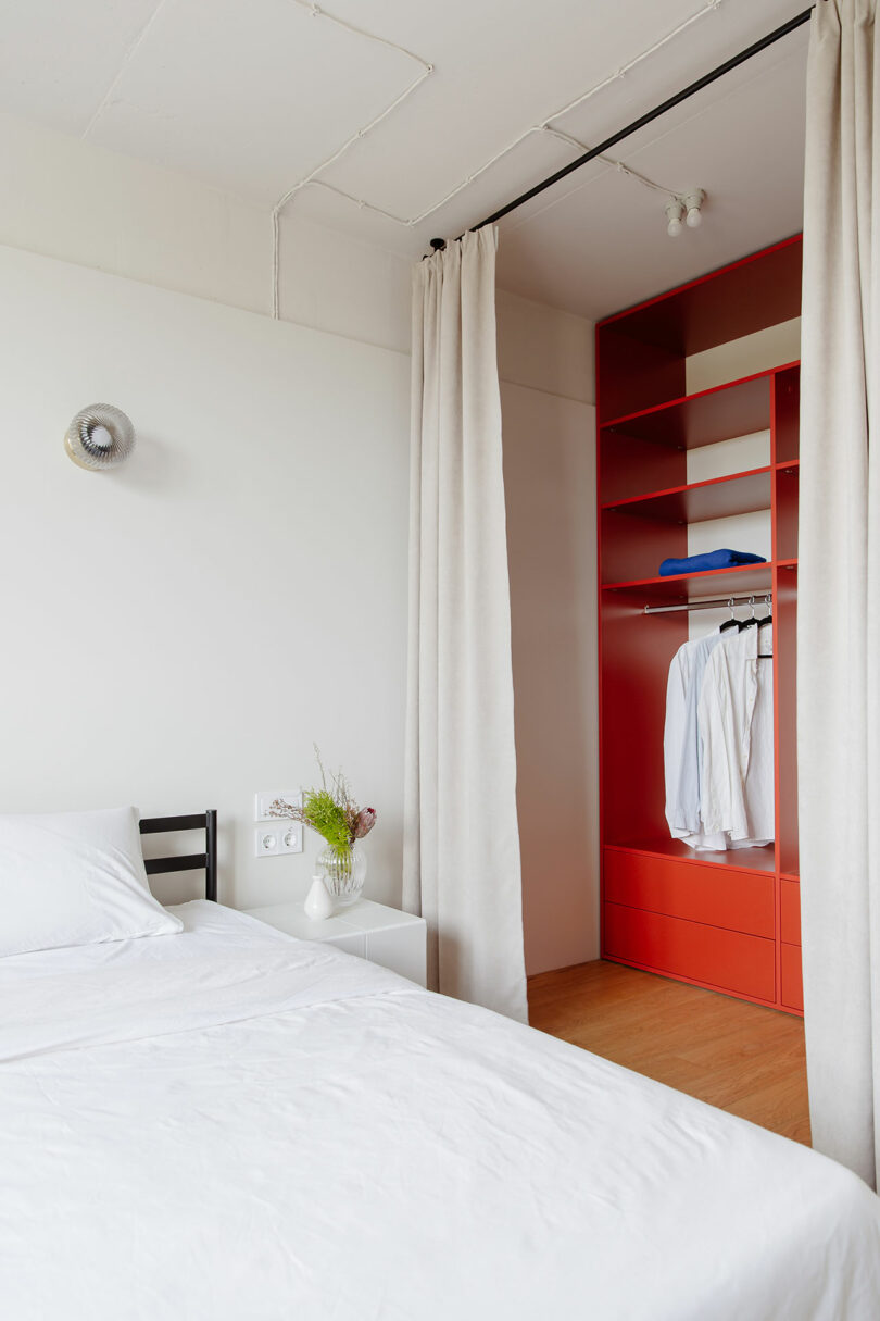 angled view of minimalist white bedroom with white bedding and curtain open to reveal bright red closet