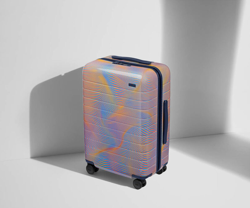 Hard-shell luggage with colorful AWAY x Resident Advisor Soundwave pattern standing against a white background with a shadow to the left.
