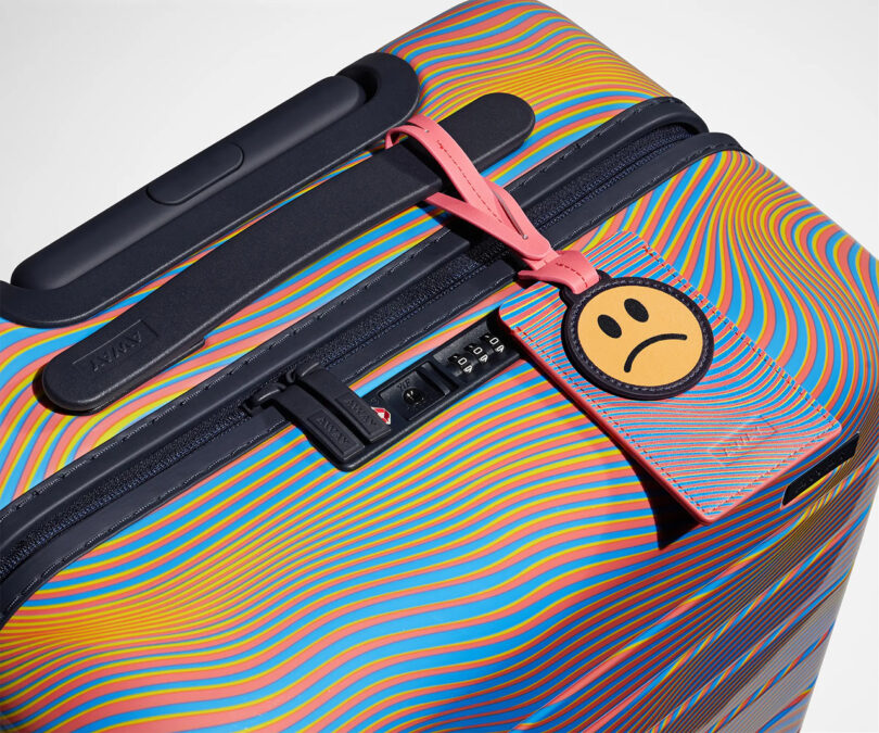 Colorful AWAY x Resident Advisor Soundwave hard-shell luggage with a combination lock and a distinctive frowning face luggage tag.