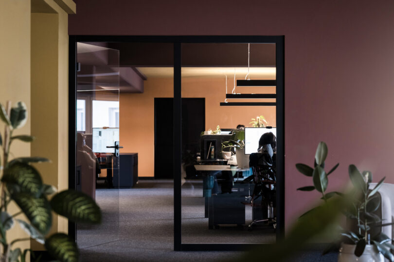A view into a modern office space through a glass doorway, with a person working at a desk surrounded by green plants and NOA.