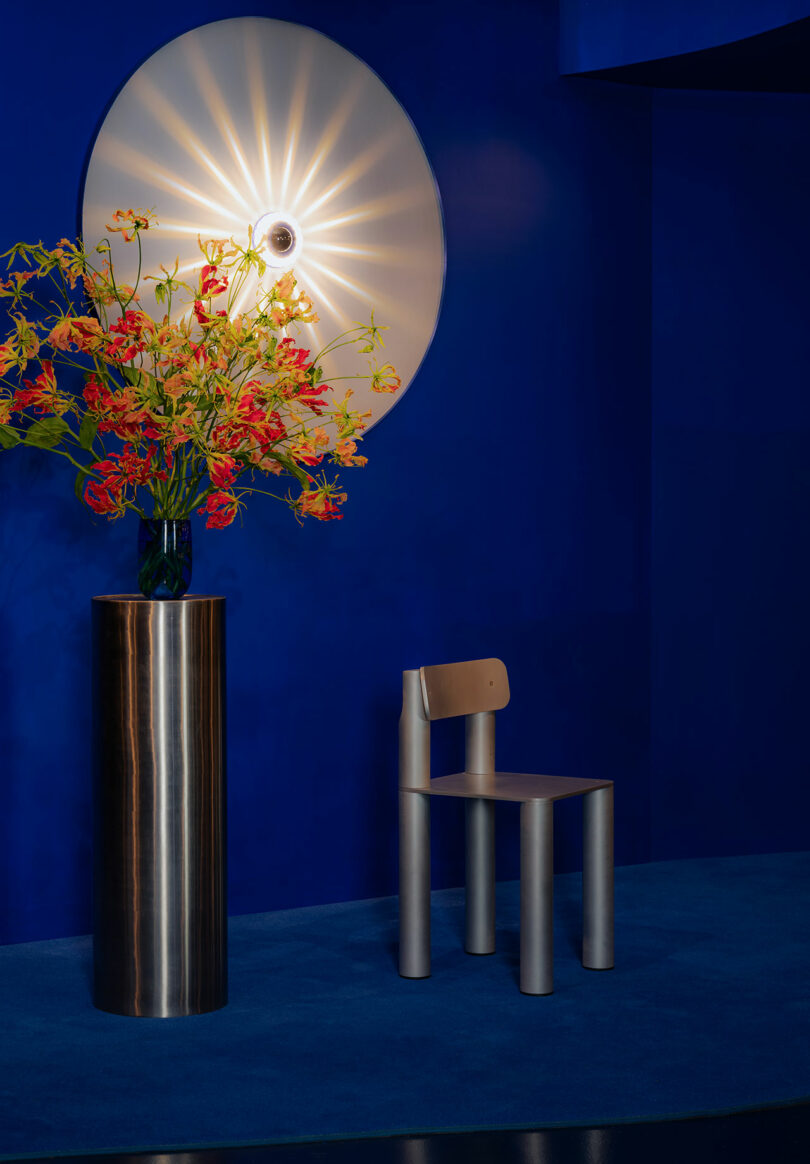 A modern interior featuring a metallic chair and a tall vase with vibrant flowers, illuminated by a large circular wall light on a deep blue background.