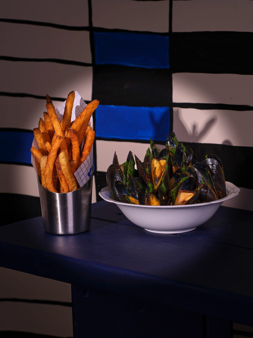 A bowl of mussels and a cup of french fries on a dark blue table against a striped blue and black wall, exuding an air of mystery that only strangers might love.