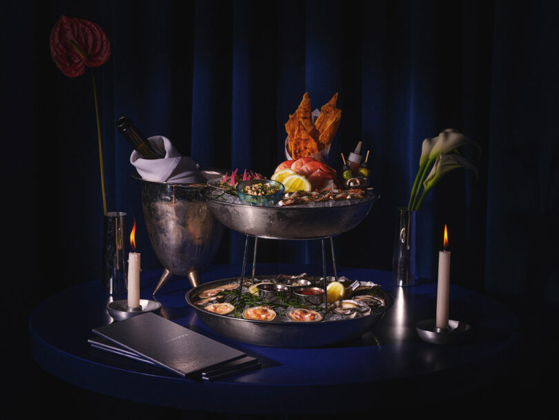 Elegant seafood dinner setup featuring a central bowl with shellfish, adjacent smaller dish with assorted seafood, candles, flowers, and dark blue ambiance designed to make strangers fall in love.