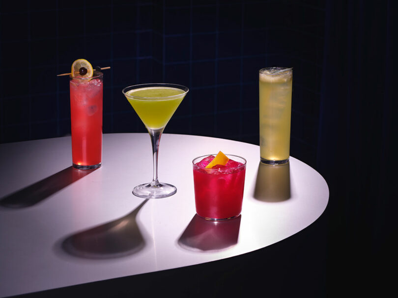 Four cocktails of varying colors presented on a round table under spotlight, creating dramatic shadows on a dark background, only enhancing the allure for strangers.