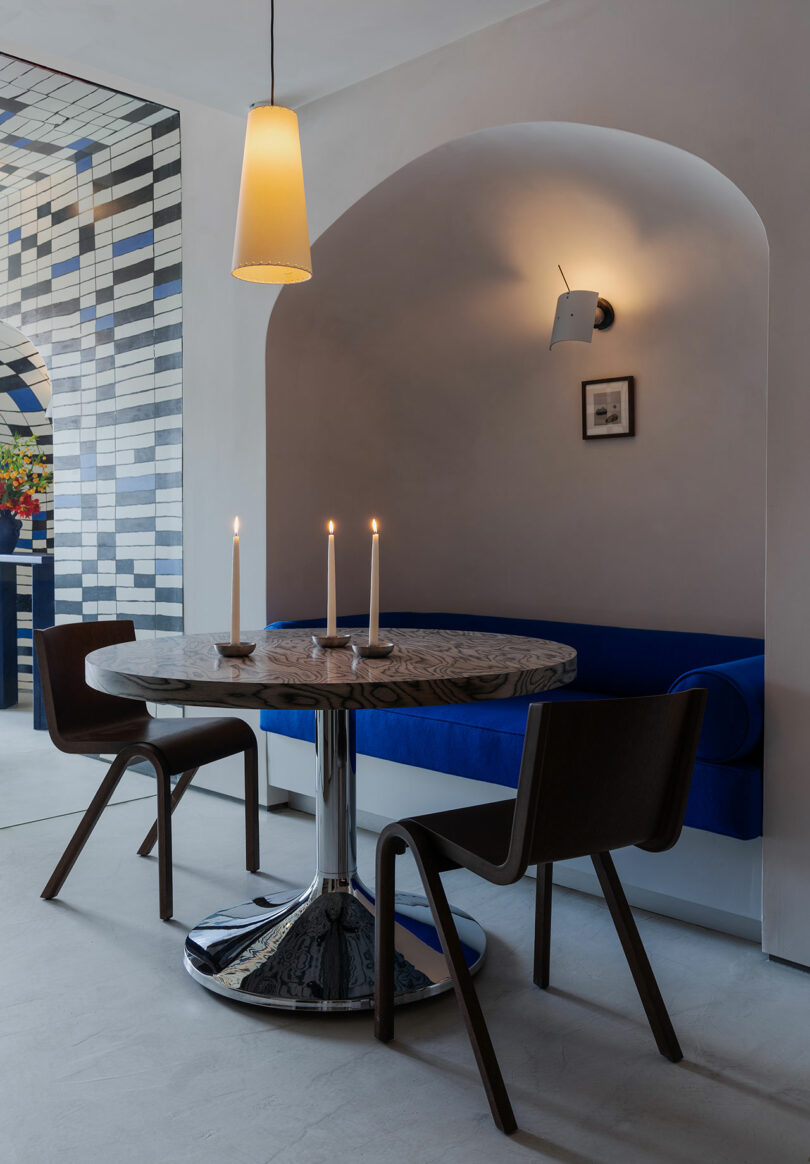 A modern dining area featuring a round marble table with candles, two chairs, a blue curved booth, white walls, and a geometric patterned wall in the background.