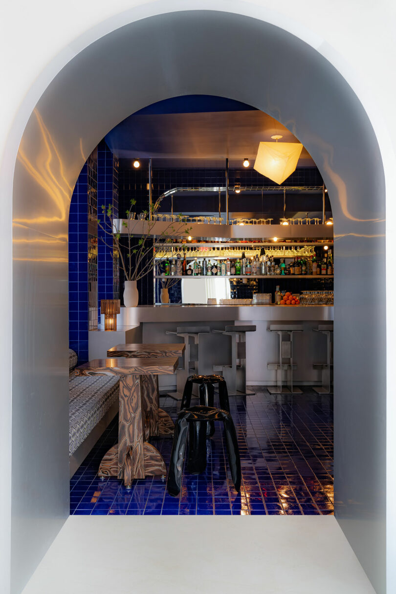 View through an arched doorway of a modern bar with blue tiles, hanging pendant light, and a well-stocked bar counter.