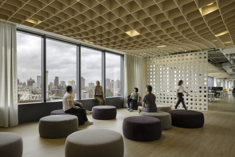 Modern office lounge area with employees sitting on round ottomans, large windows showcasing city skyline, and unique wooden ceiling design