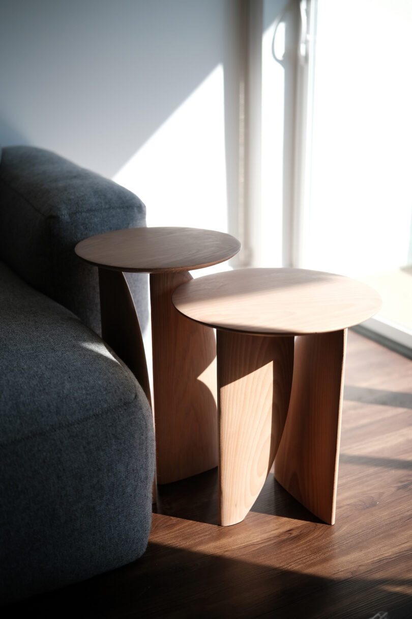 Two nested wooden side tables next to a gray sofa in a sunlit room with a hardwood floor and a glimpse of a window.