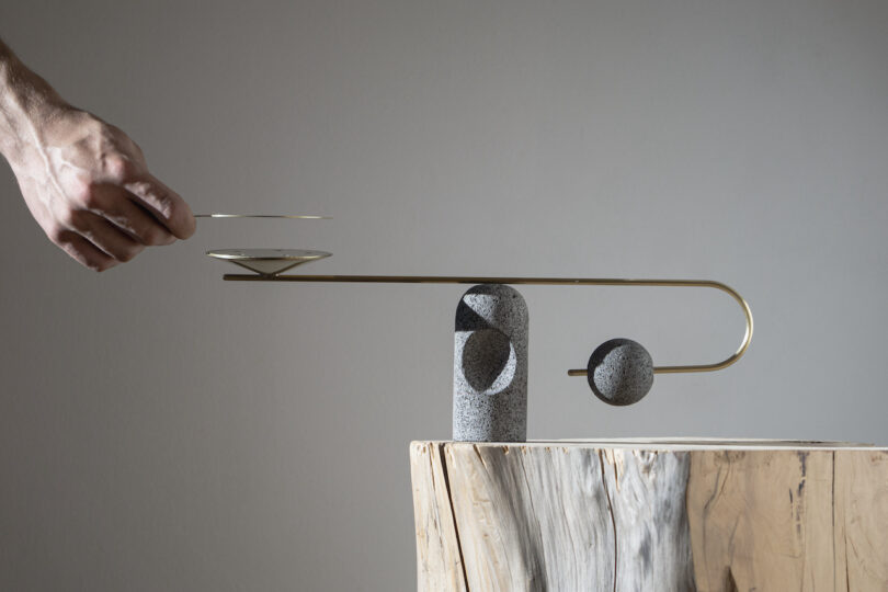 A hand placing a saucer on a minimalist, balance-inspired sculpture next to a section of polished marble.