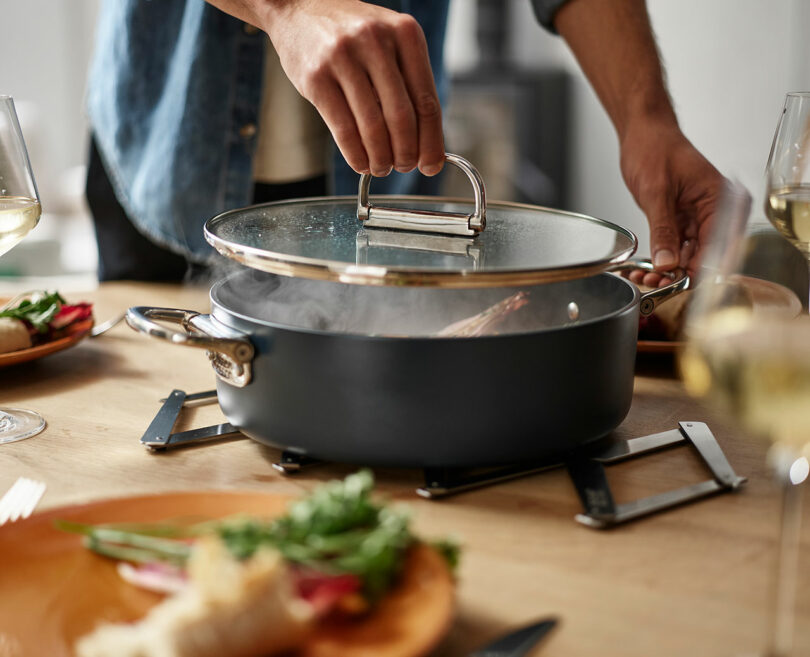 A person lifting the lid of a steaming pot on a dining table while preparing to serve food.