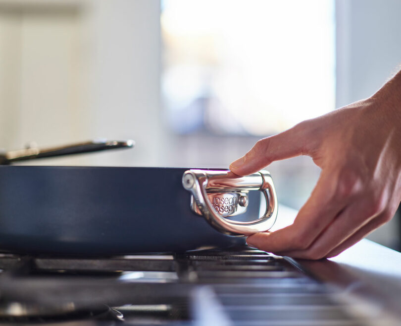 Person's hand holding a pot lid handle over a stove-top.