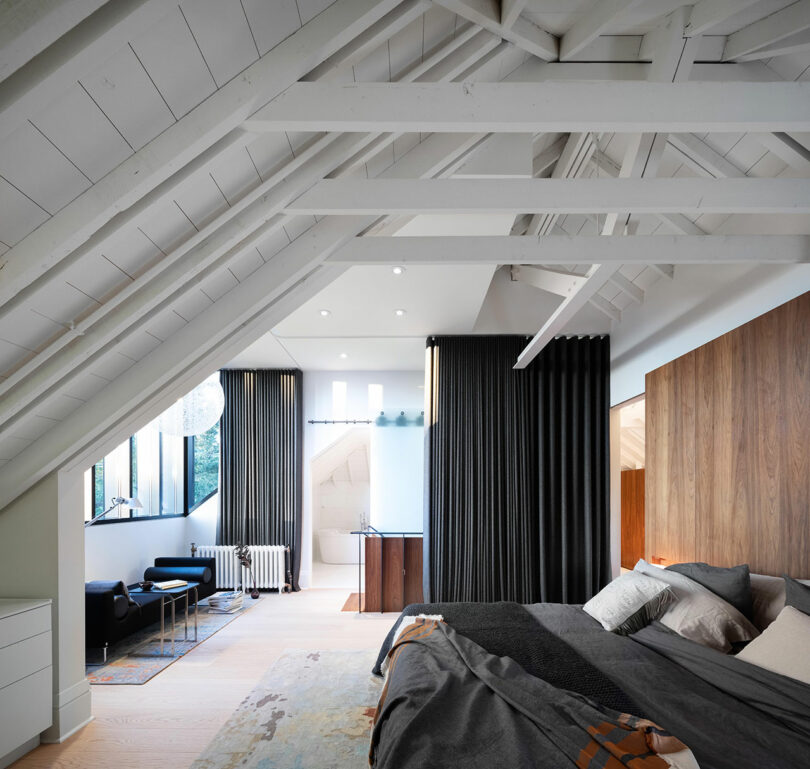 Modern attic bedroom with exposed white beams, a wooden wall panel, large bed, and black curtains dividing the space.