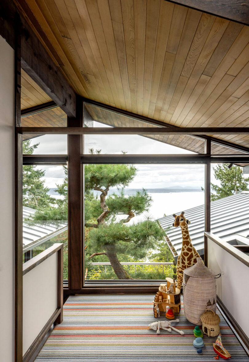 A bright, airy corridor with large windows overlooking a tree and the sea, featuring a toy giraffe and colorful striped rug.